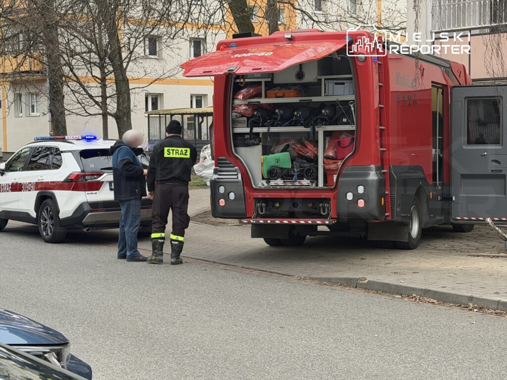 Mężczyzna w niebieskiej kurtce rozmawia z ratownikiem w mundurze przy wozie strażackim na ulicy.