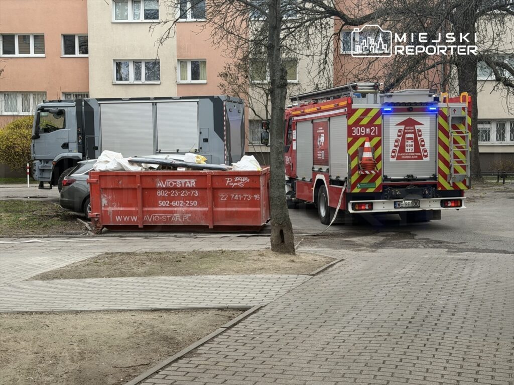 Straż pożarna podjeżdża do czerwonego kontenera na odpady w otoczeniu bloków mieszkalnych.