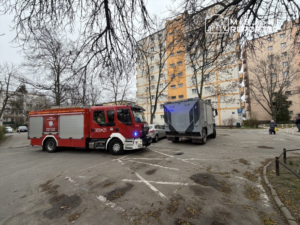 Czerwony wóz strażacki z włączonymi sygnałami stoi na parkingu obok szarego pojazdu w pobliżu bloków mieszkalnych.