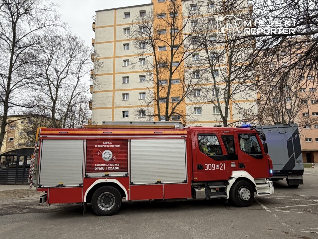 Czerwony wóz strażacki zaparkowany przed blokiem mieszkalnym, z widoczną informacją o bezpieczeństwie.