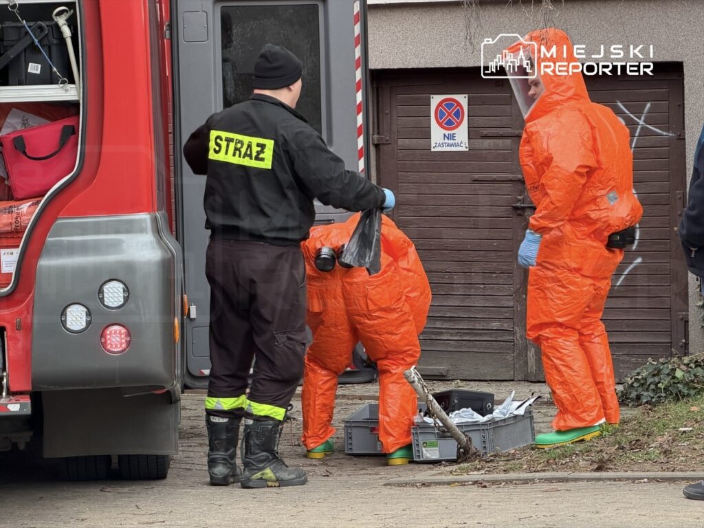 Trzech ratowników w pomarańczowych kombinezonach zabezpieczających przeszukuje teren obok wozu strażackiego.