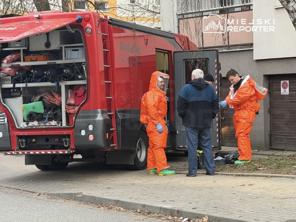 Czterej ratownicy w pomarańczowych kombinezonach rozmawiają z mężczyzną na ulicy obok pojazdu ratunkowego.