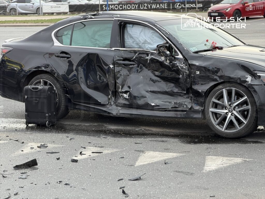 Czarny samochód osobowy z uszkodzoną lewą stroną stoi na drodze, obok walizka i odłamków szkła.