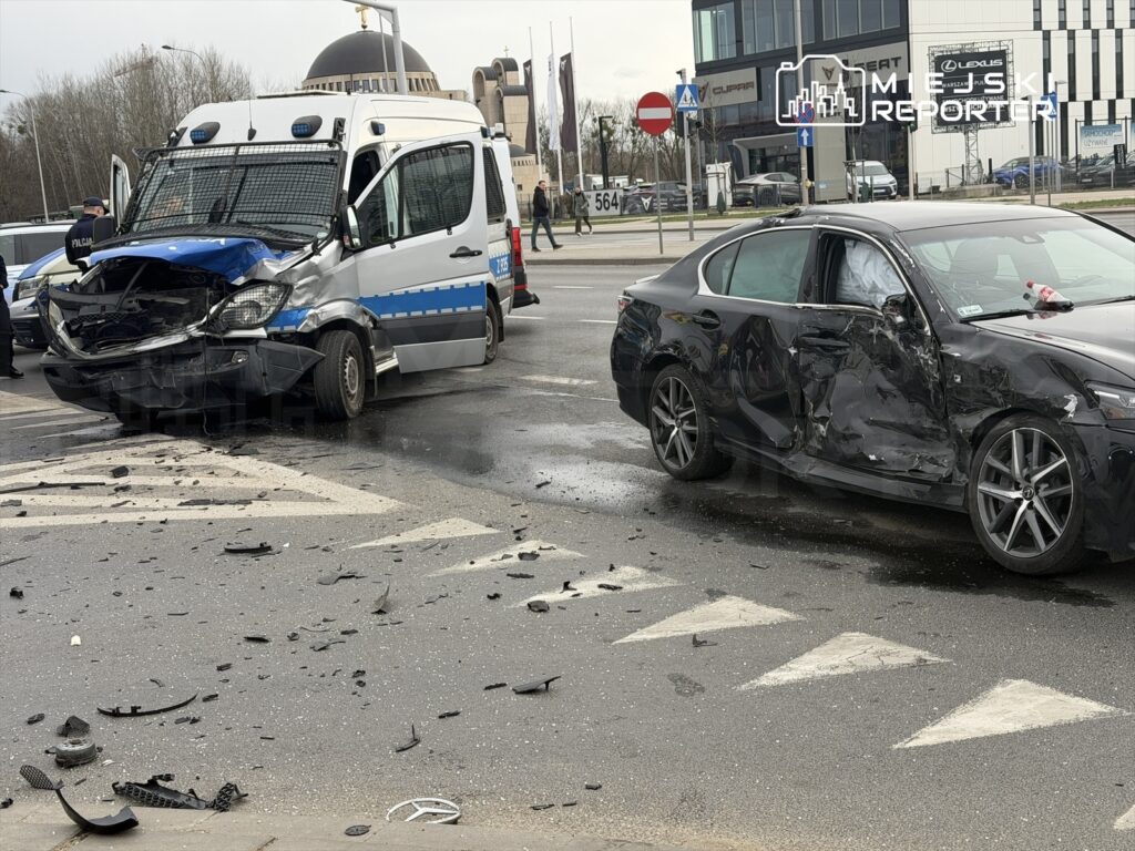 Zderzenie dwóch samochodów na skrzyżowaniu, z uszkodzonym furgonem policyjnym i czarnym autem osobowym. Na jezdni leżą fragmenty pojazdów.