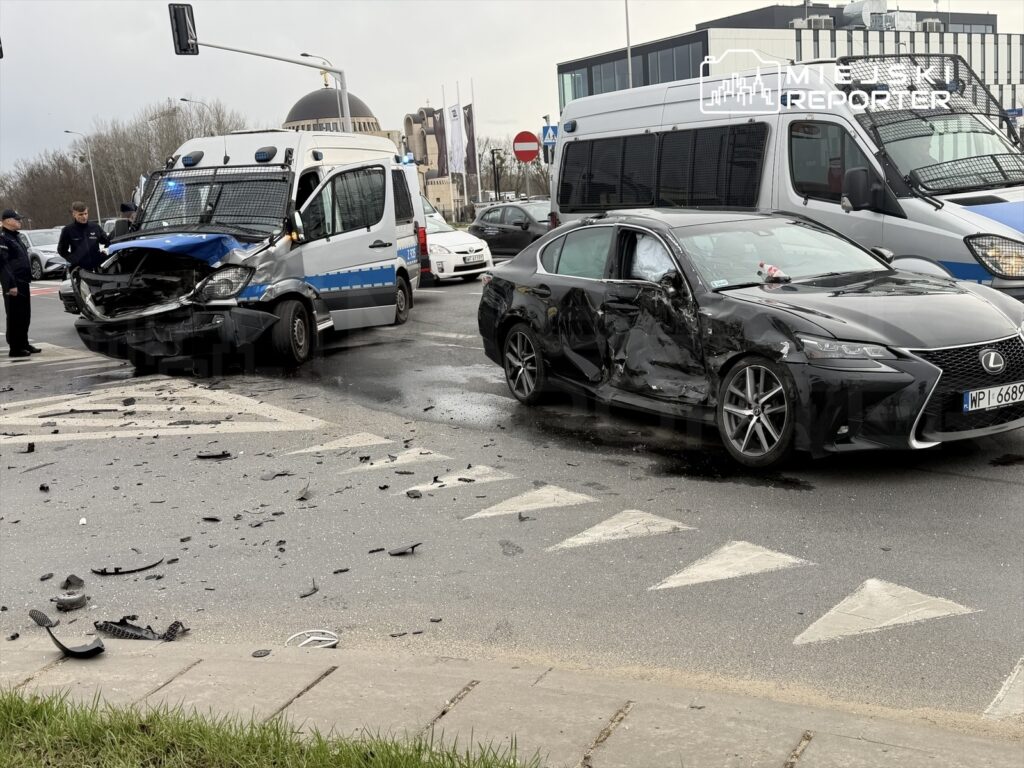 W wyniku kolizji drogowej uszkodzone pojazdy: policyjny furgon z uszkodzoną przednią częścią oraz czarny samochód osobowy z wgnieceniem na boku. Na drodze leżą fragmenty karoserii.
