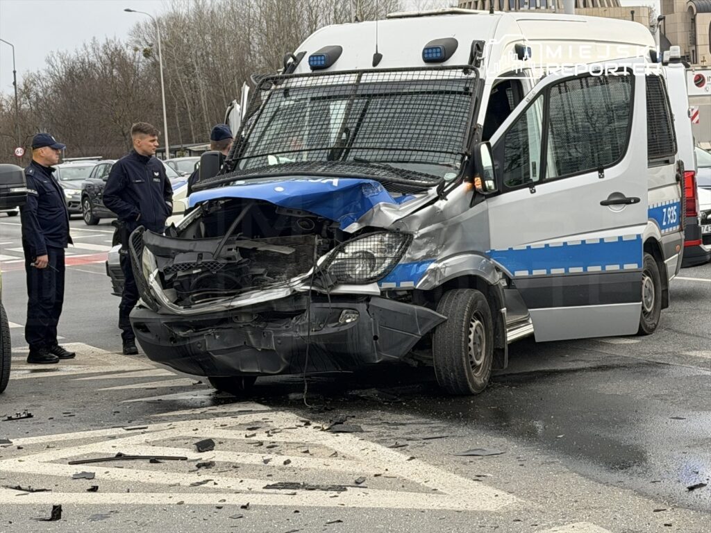 Policyjny furgon z uszkodzoną przednią częścią stoi na skrzyżowaniu, obok trzech funkcjonariuszy rozmawiających o wypadku.