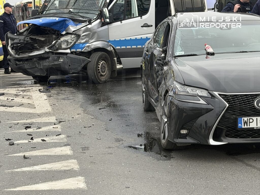 Zniszczony furgon policyjny i uszkodzony czarny samochód osobowy na ruchliwej ulicy, wokół rozbite elementy karoserii.