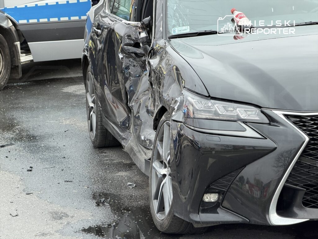 Uszkodzony czarny samochód osobowy z wgniecioną lewą stroną stoi na mokrej nawierzchni, obok policyjnego radiowozu.