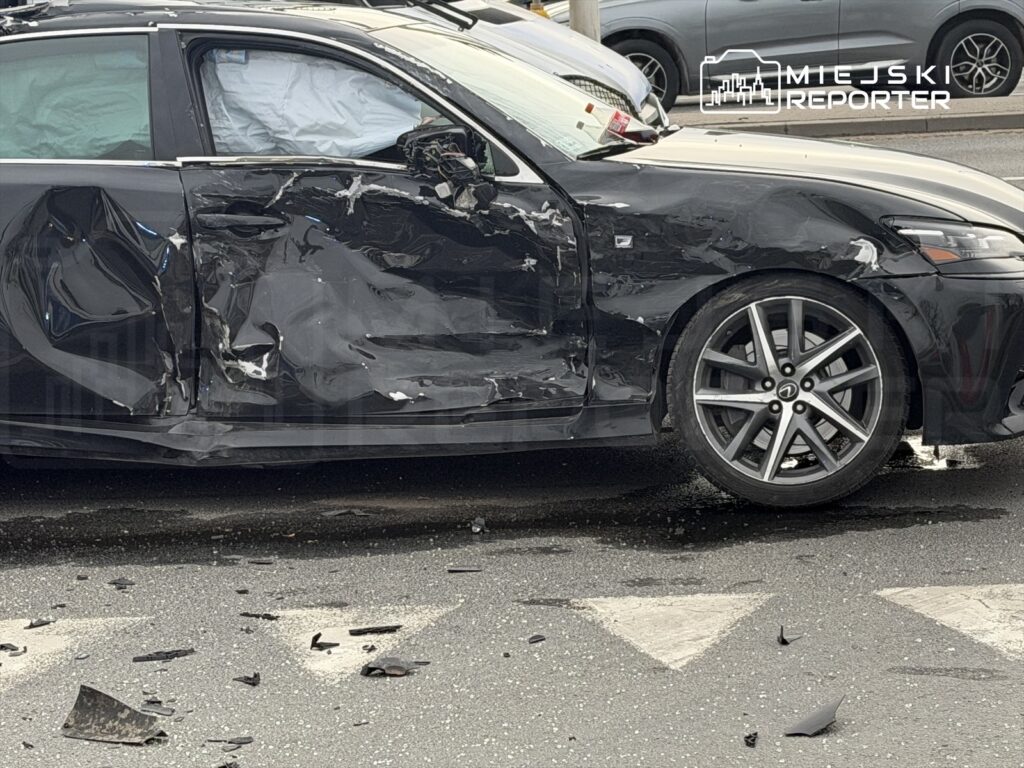 Uszkodzony czarny samochód osobowy z wgniecioną boczną częścią stoi na drodze, wokół leżą fragmenty karoserii.