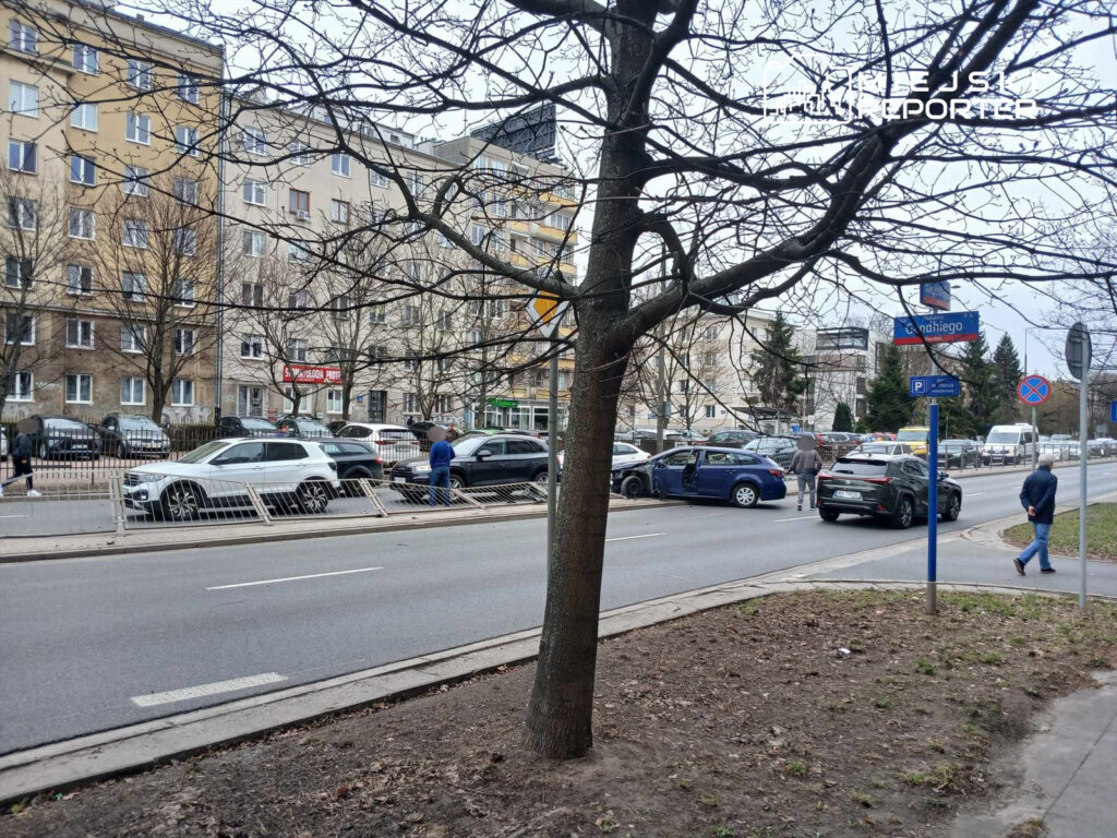 Na ulicy zaparkowane samochody, w tle mężczyzna rozmawiający z innym przy uszkodzonym pojeździe.