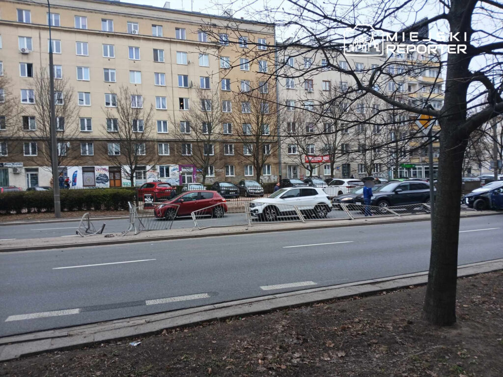 Ruchliwa ulica z samochodami, na pierwszym planie barierki, a w tle budynek mieszkalny z oknami.