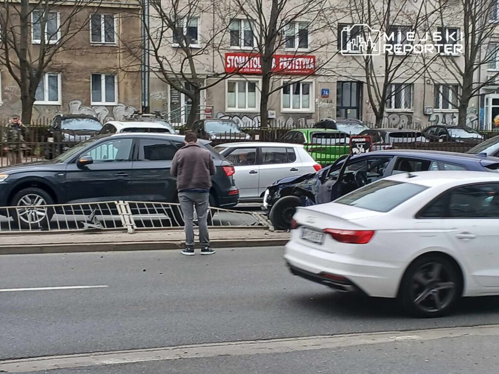 Mężczyzna w szarej bluzie stoi obok uszkodzonego samochodu na ulicy, obok przejeżdżających pojazdów.