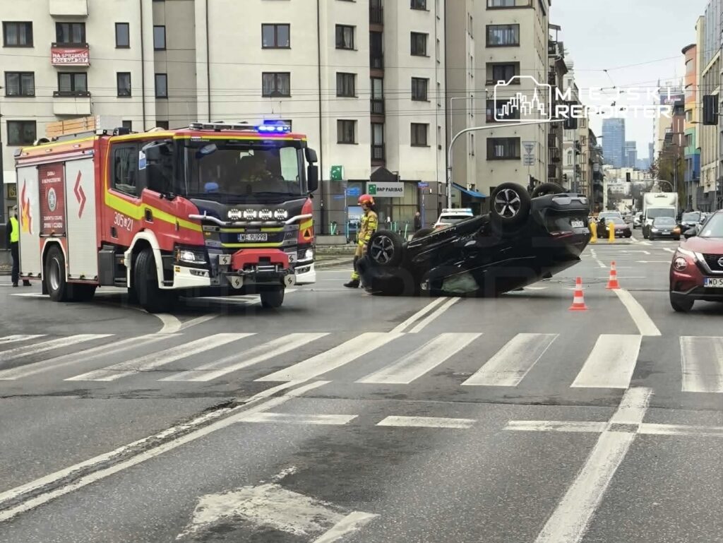 Na ulicy leży przewrócony na dach czarny samochód, obok niego stoi wóz strażacki z włączonymi sygnałami świetlnymi.