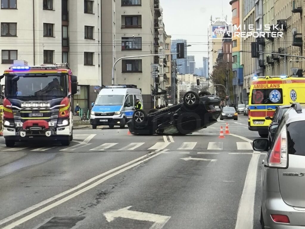 Na ulicy leży przewrócony samochód, obok stoją pojazdy straży pożarnej i karetka, a w tle widać budynki.