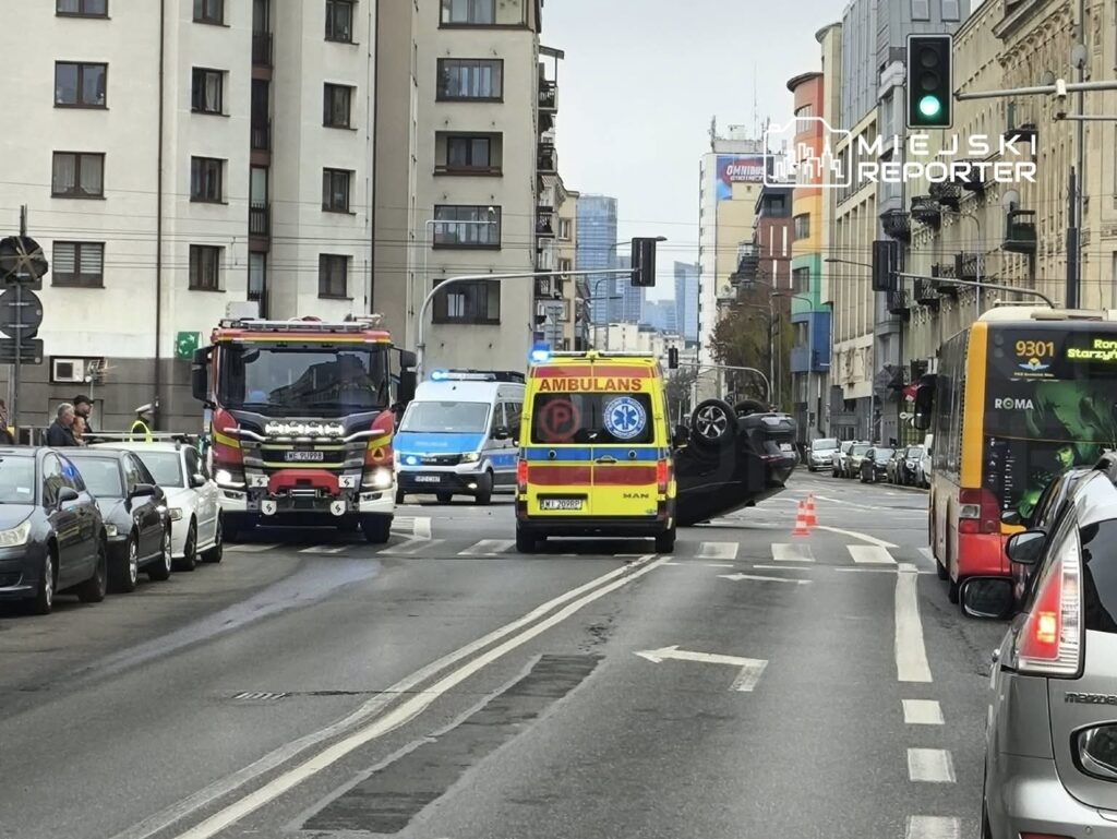 Na ulicy w mieście, samochód osobowy leży na dachu, a obok znajdują się pojazdy służb ratunkowych: straż pożarna i karetka.
