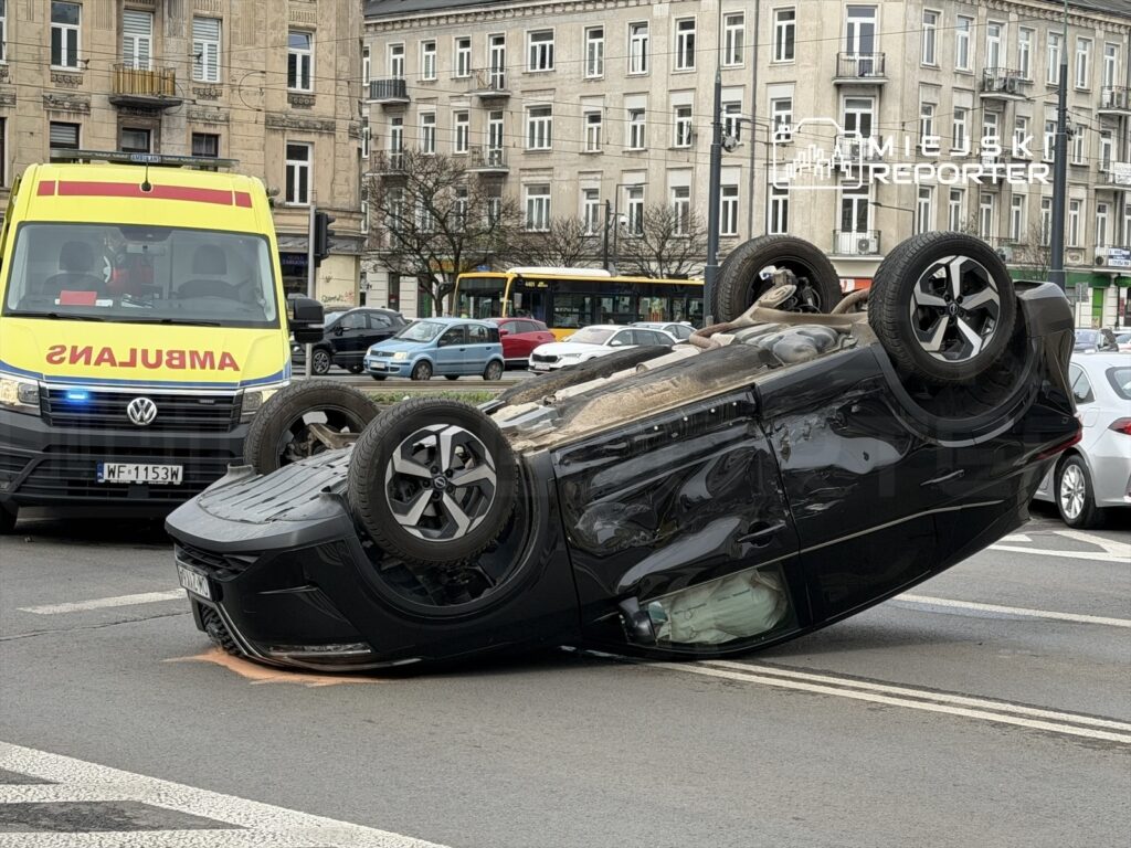 Czarny samochód osobowy leży na dachu na jezdni, obok stoi ambulans z włączonymi sygnałami świetlnymi.