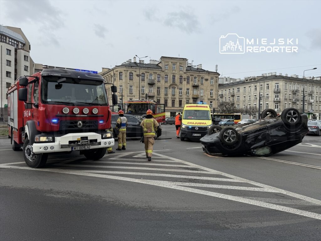 Czarny samochód leży na dachu na skrzyżowaniu, obok strażacy i karetka pogotowia w akcji.