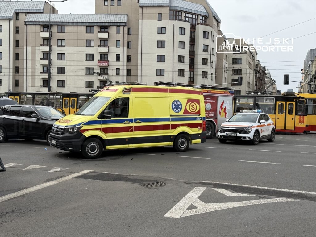 Karetka pogotowia w żółto-czerwonym kolorze oraz radiowóz policji zatrzymane na skrzyżowaniu w miejskim otoczeniu.