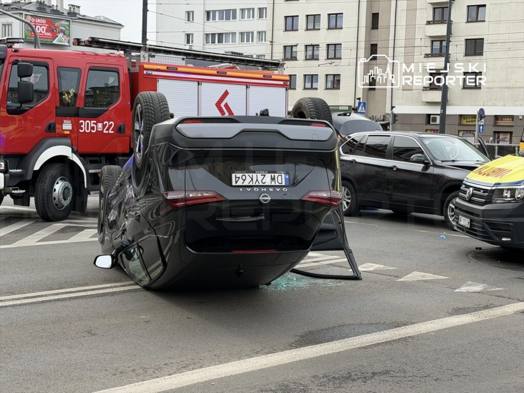 Czarny samochód osobowy leży na dachu na ruchliwej ulicy, obok wozu strażackiego i innych pojazdów.