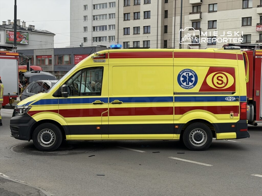 Żółto-czerwony ambulans z oznaczeniem "Ratownictwo Medyczne" stoi na ulicy obok wozu strażackiego.