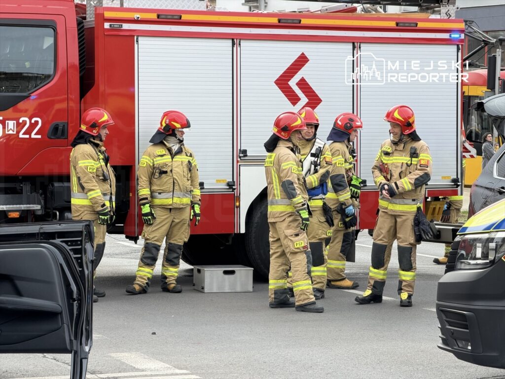 Grupa strażaków w mundurach rozmawia obok wozu strażackiego na miejskiej ulicy.