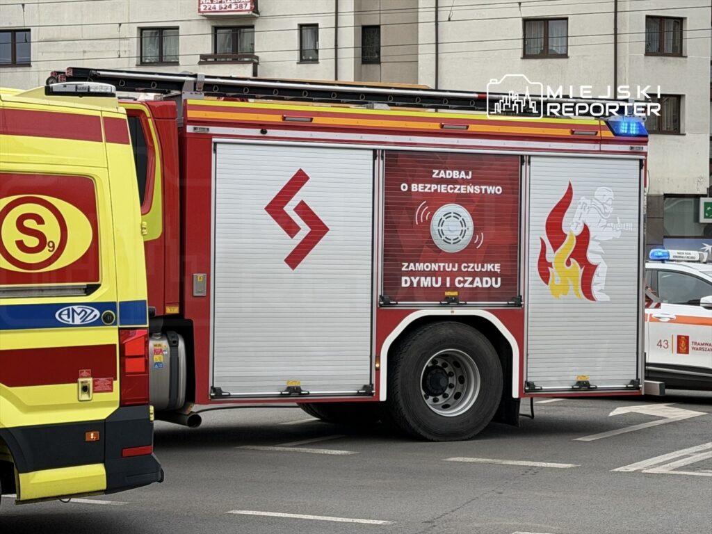 Czerwony wóz strażacki z grafiką ostrzegawczą stoi na skrzyżowaniu, obok żółtego pojazdu ratunkowego.