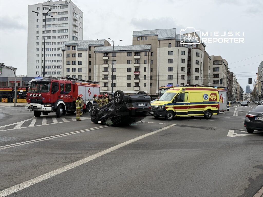 Na ulicy leży przewrócony samochód, obok niego strażacy oraz karetka pogotowia. W tle widać budynki mieszkalne.
