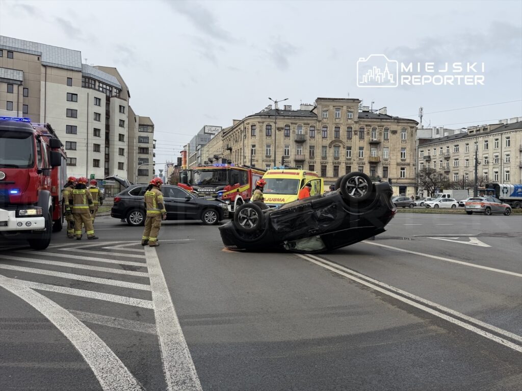 Na skrzyżowaniu przewrócone na dach czarne auto, wokół strażacy w mundurach i pojazdy ratunkowe.