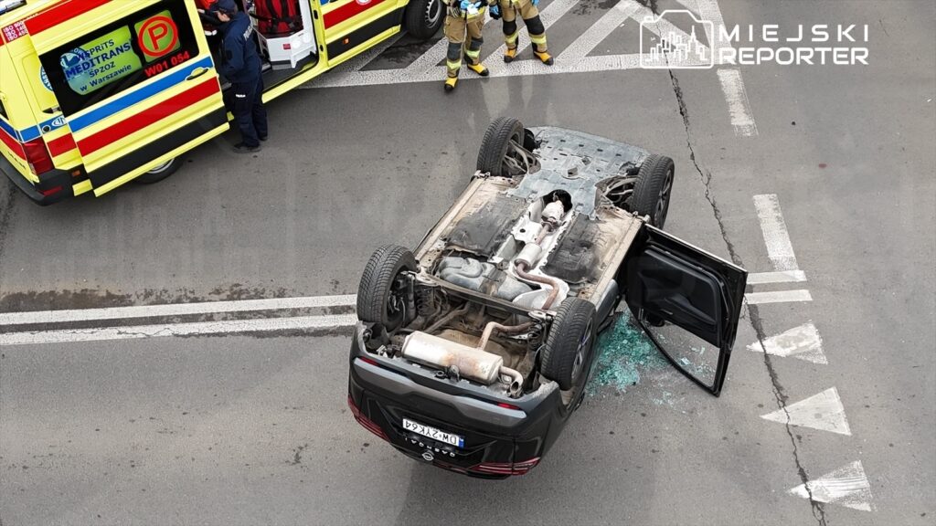 Odwrócone auto leży na drodze, wokół niego pracują strażacy i ratownicy medyczni, a na jezdni widać rozbite szkło.