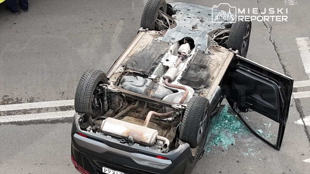 Odwrócone auto leży na jezdni, z uszkodzoną szybą i otwartymi drzwiami, obok widoczne fragmenty szkła.