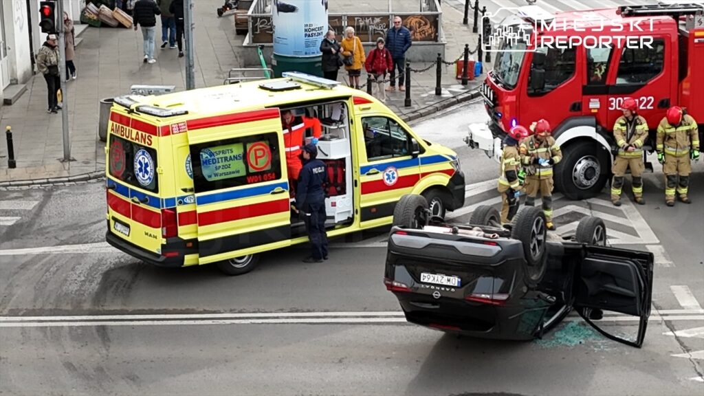 Karetka pogotowia i strażacy pracują na miejscu wypadku drogowego, gdzie przewrócone auto leży na dachu.