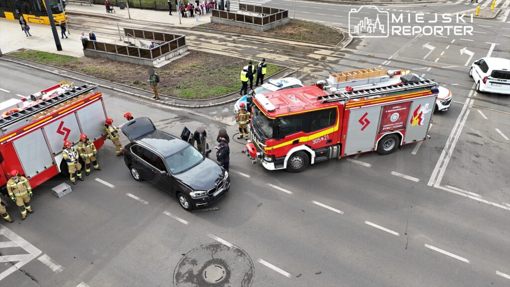 Grupa strażaków w mundurach dokonuje oględzin uszkodzonego czarnego SUV-a przy włączonym wozie strażackim na skrzyżowaniu.