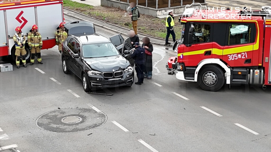 Czterech strażaków w mundurach stoi obok uszkodzonego czarnego SUV-a na drodze, w tle widać wóz strażacki.