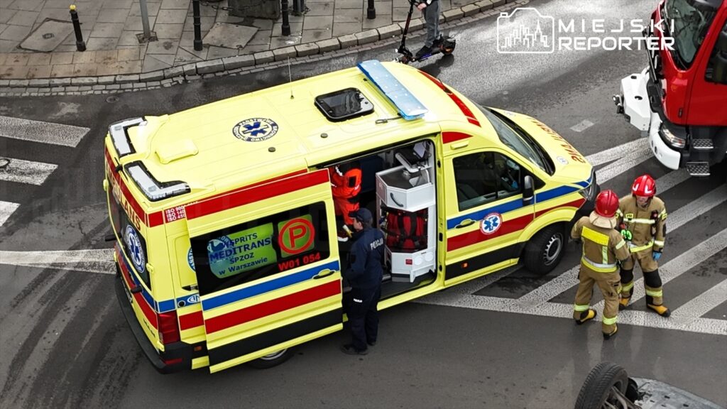 Ambulans z otwartymi drzwiami, z zespołem ratunkowym w pomarańczowych uniformach, rozmawia z policjantem na skrzyżowaniu.