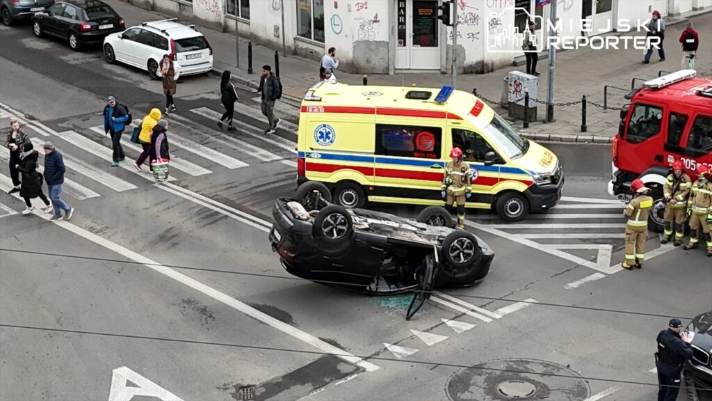 Na skrzyżowaniu leży przewrócone na dach czarne auto, obok niego stoją ratownicy medyczni i strażacy. W tle widać przechodniów oraz karetkę pogotowia.