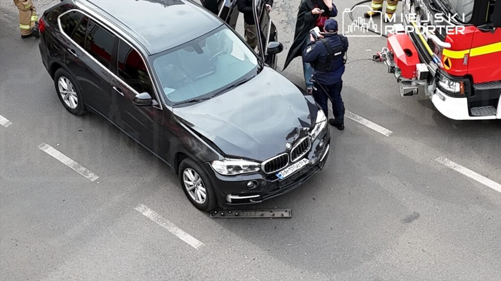 Czarny SUV stoi na drodze, obok grupka osób w mundurach rozmawia, a w tle widoczny wóz strażacki.