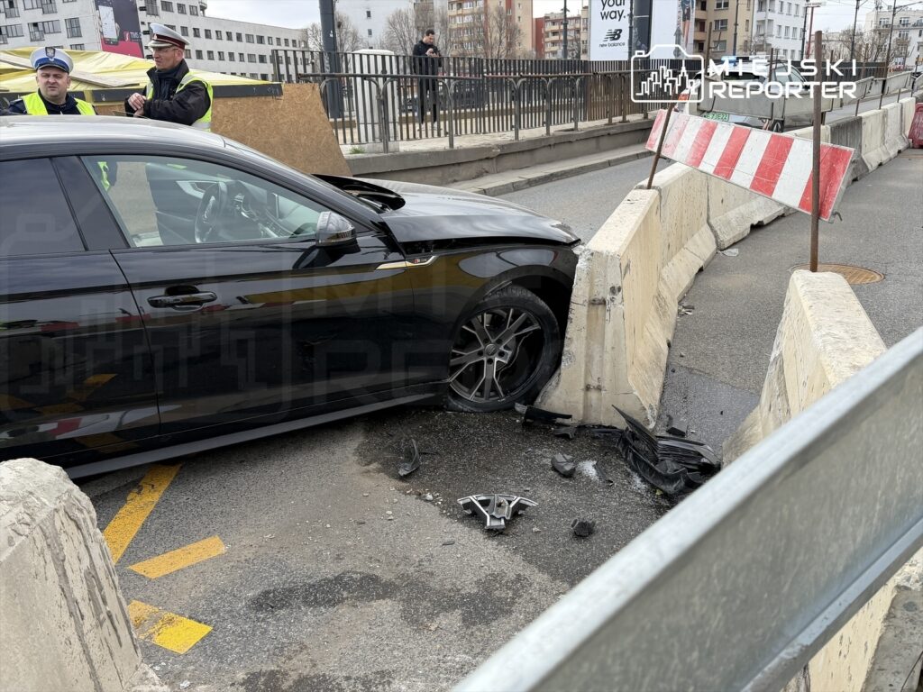 Czarny samochód osobowy wjeżdżający w betonowy blok na jezdni, obok dwóch policjantów w odblaskowych kamizelkach.