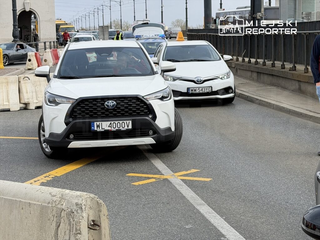 Biały samochód osobowy z widocznym numerem rejestracyjnym blokuje ruch na ulicy, otoczony przez inne pojazdy.