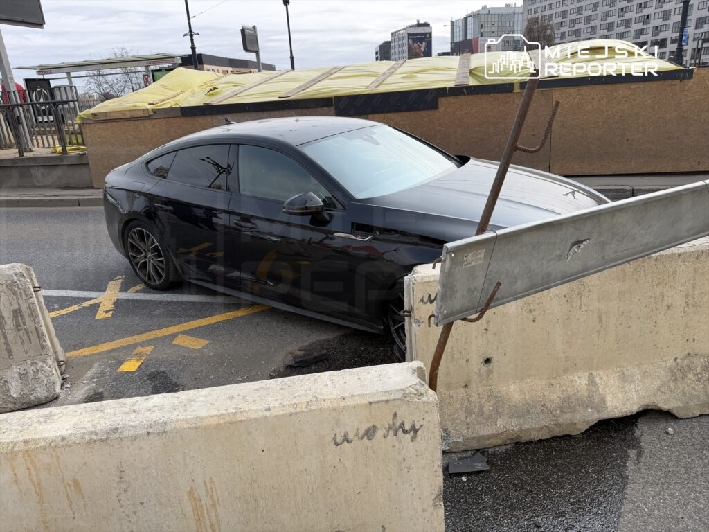 Czarny samochód osobowy utknął pomiędzy betonowymi blokadami na ruchliwej ulicy.