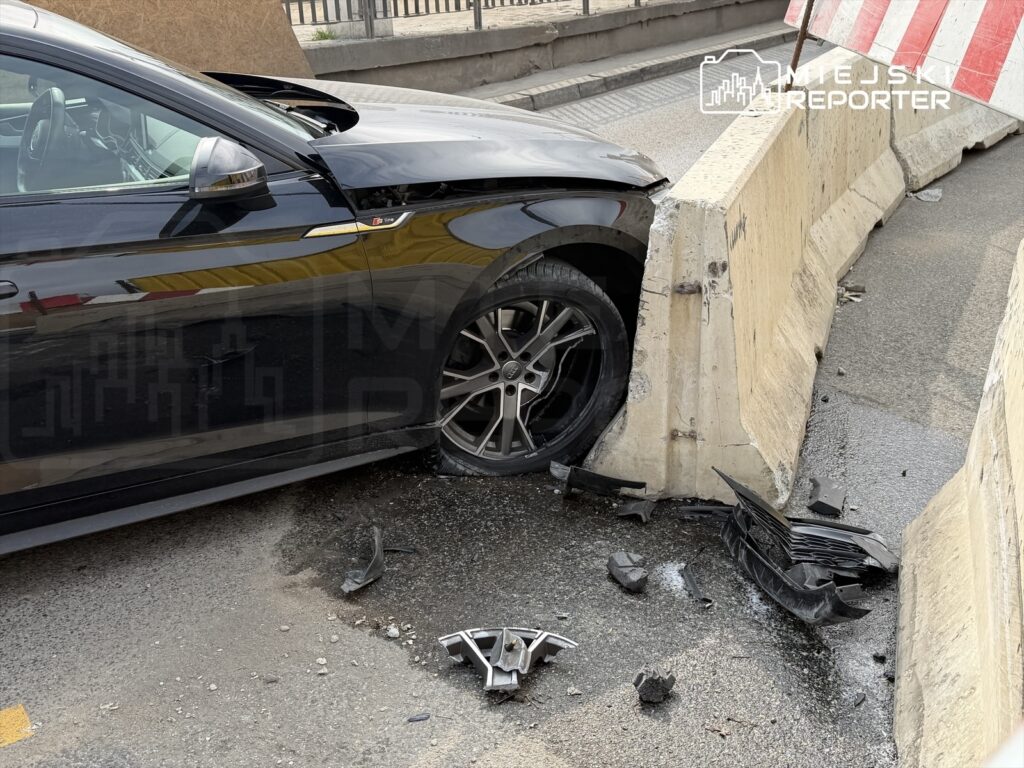 Czarny samochód osobowy uderzył w betonowy separator na jezdni, obok leżą fragmenty zderzaka.