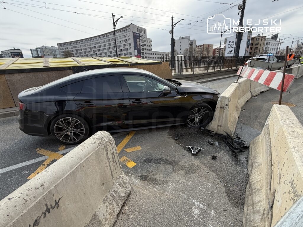 Czarny samochód osobowy uderzył w betonową barierę na drodze, obok leżą resztki zderzaka. W tle widać budynki.