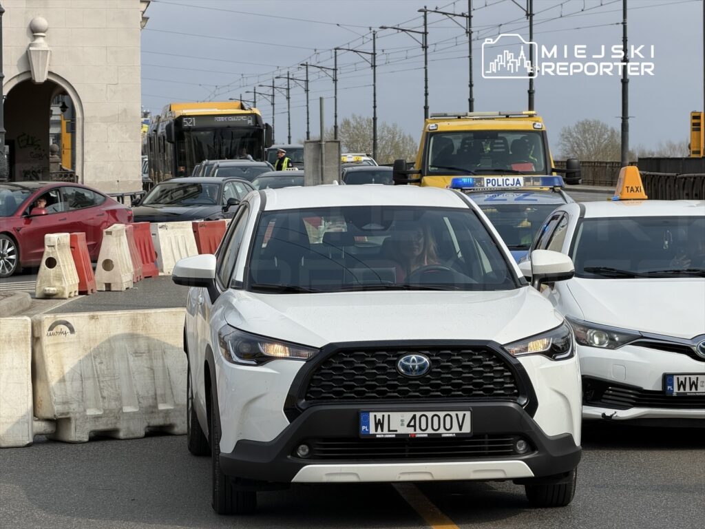 Biały samochód osobowy stoi w zatorze drogowym, a w tle widoczne są pojazdy komunikacji miejskiej i radiowóz policyjny.