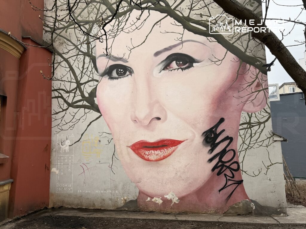 Murale przedstawiające dużą twarz kobiety z czerwonymi ustami, z gałęziami drzew w tle oraz graffiti na policzku.