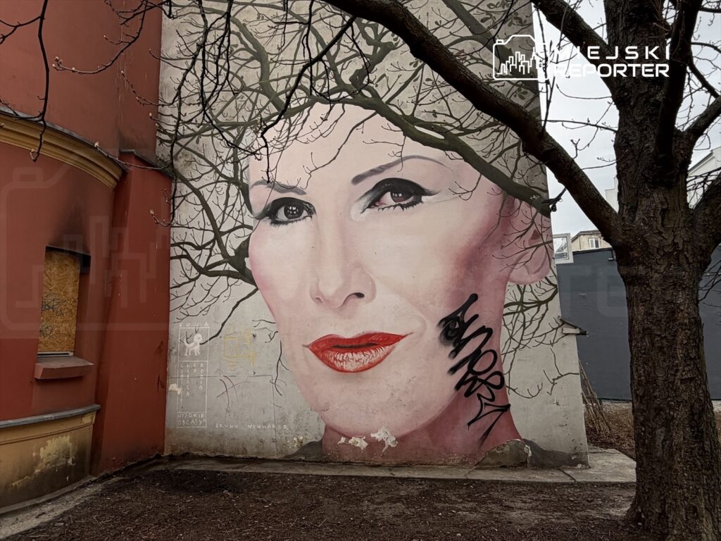 Murale przedstawiające kobiecą twarz z czerwonymi ustami, otoczone gałęziami drzew, na ścianie budynku.