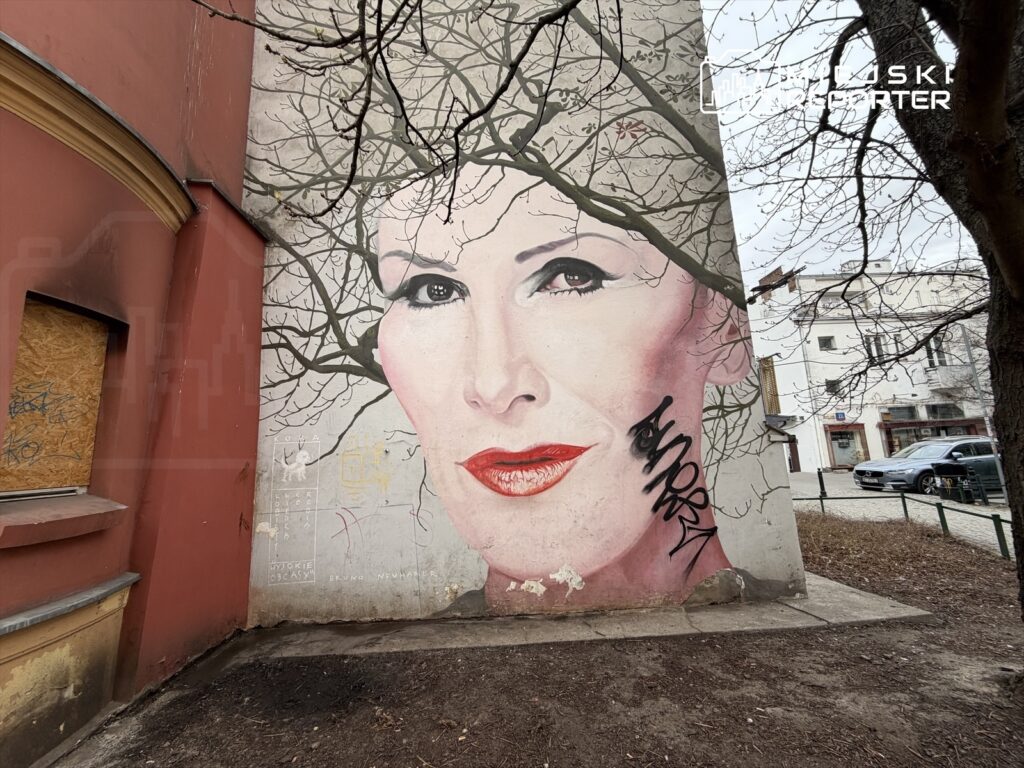 Mural przedstawiający dużą twarz kobiety z czerwonymi ustami, otoczoną gałęziami drzew, na ścianie budynku.