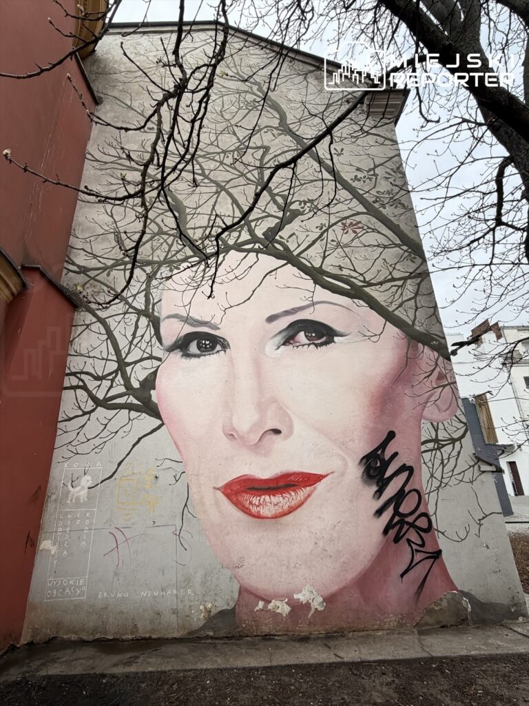 Mural przedstawiający twarz kobiety z czerwonymi ustami, otoczoną gałęziami drzew, na ścianie budynku.