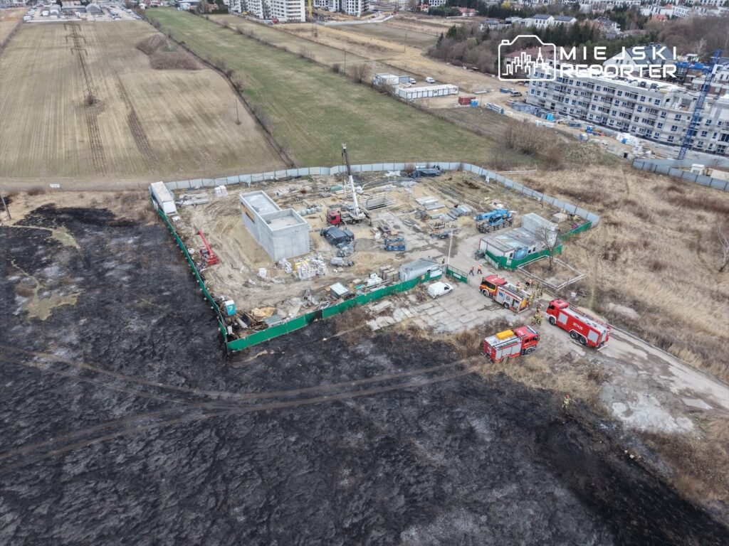 Na placu budowy z betonowym fundamentem pracują maszyny, a w tle widać straż pożarną i spalone pole.