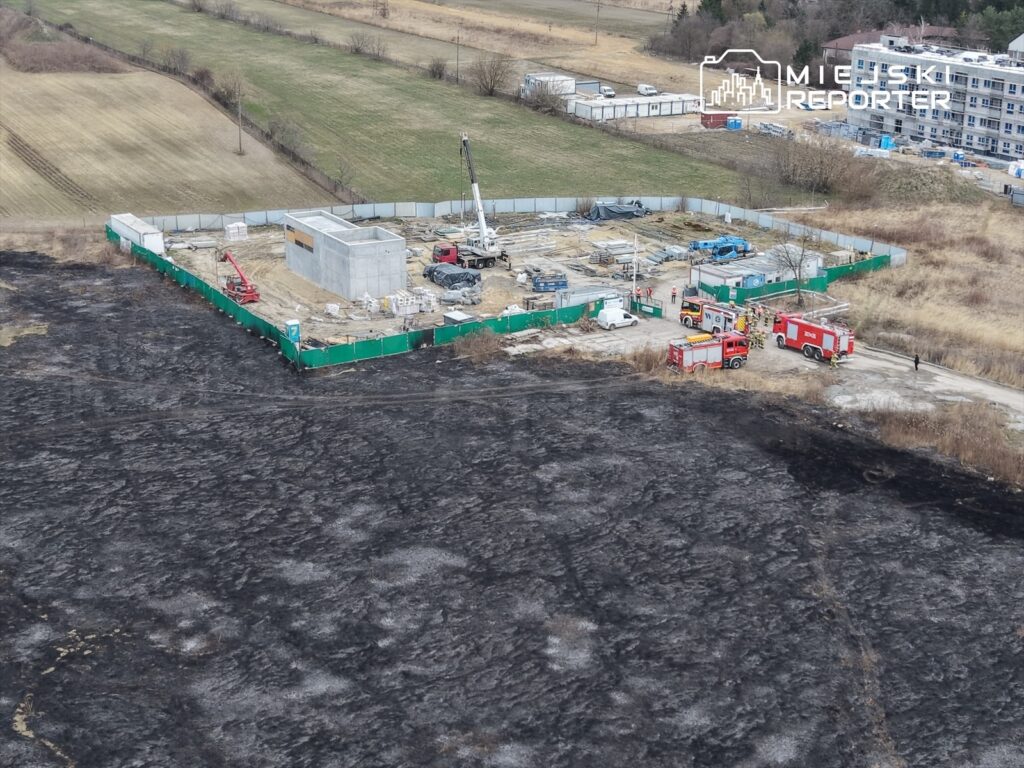 Na placu budowy widać betonowy budynek oraz sprzęt budowlany, w tle strażacy i pojazdy ratunkowe. Obszar wokół jest poparzony.