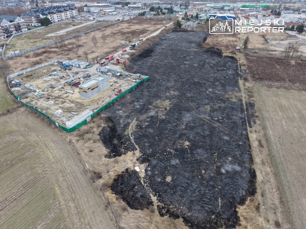 Na zdjęciu widoczny jest teren budowy z nowym budynkiem w trakcie realizacji, otoczony przez spalone pola.