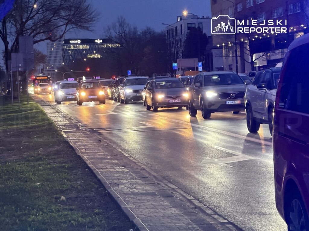 Ruch samochodowy na oświetlonej ulicy w mieście, z widocznymi światłami pojazdów i budynkami w tle.